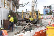 A drilling rig in an urban area with three men in high visibility gear standing around it