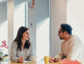 At a table in a work office, a woman with a pen and notebook talks to a man