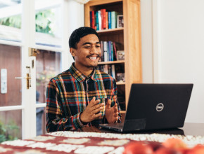 A man sits at a laptop on a table. He's smiling and clapping his hands