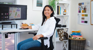 Nicole Sos is facing the camera but sitting at a desk in front of a computer in an office 
