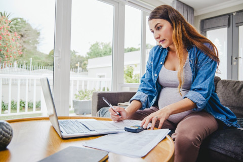 A pregnant person sits at a low table in a house looking at a laptop, while using a calculator 