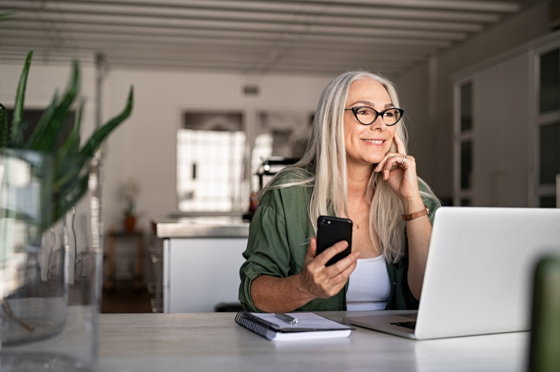 A woman sits at a desk smiling. In her hand is a mobile phone, in front of her is a laptop. She is working.