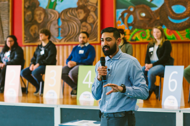 A man holding a microphone stands in front of a panel of people in a New Zealand school hall