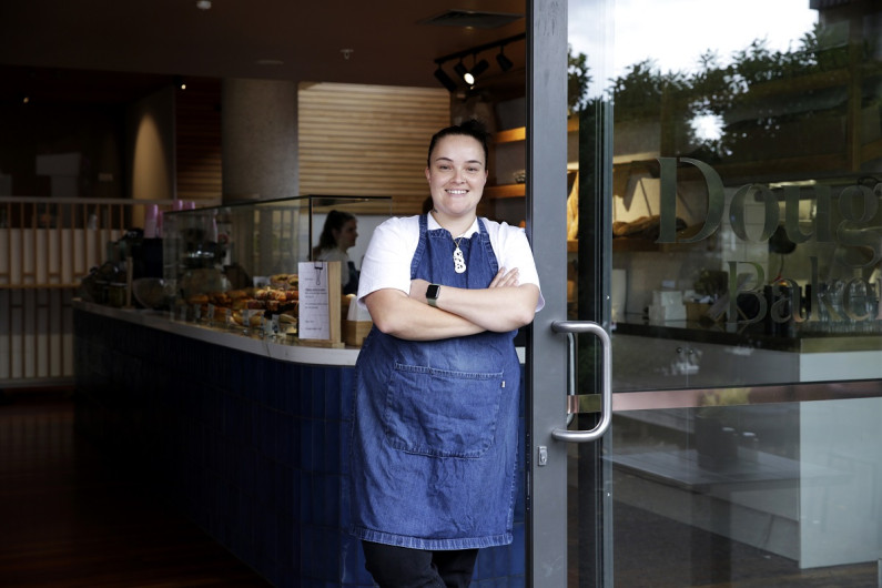 Emma Utanga stands at the front of a cafe smiling and wearing an apron with her arms folded 