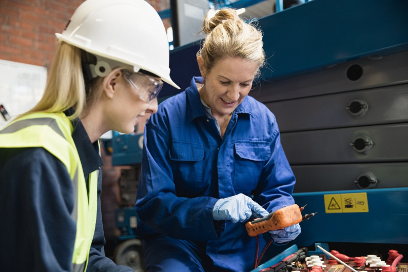 In a workshop. An employer dressed in overalls shows a high school learner in a hard hat a piece of equipment.