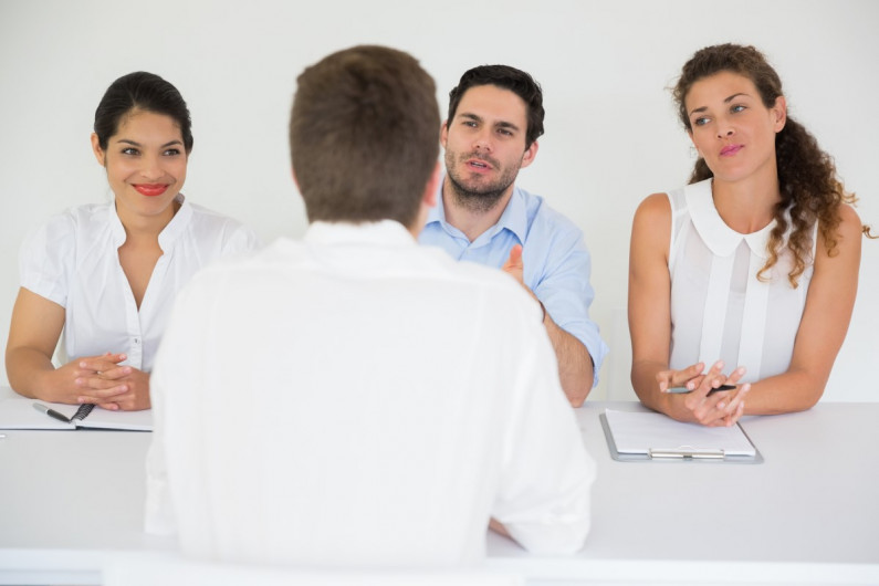 Two women and a man sitting at a table interviewing a man opposite them