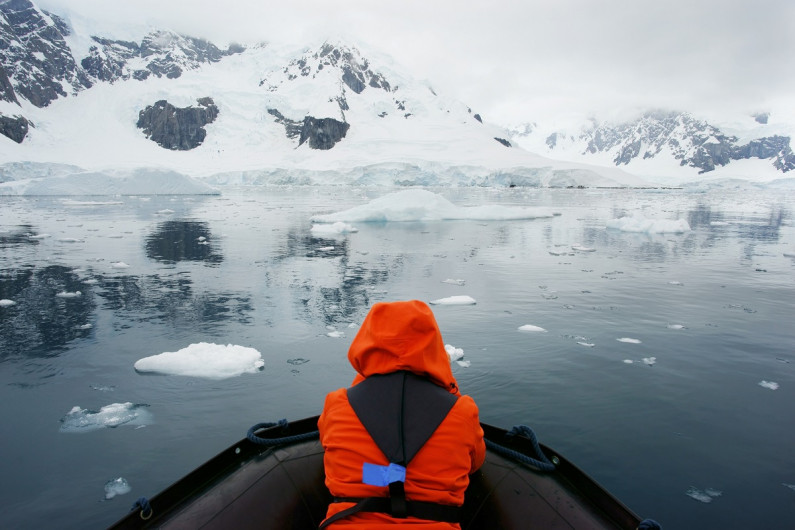 A person dressed in warm gear sits on the front of the boat looking at snow covered hills and icebergs.