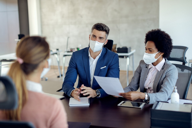 A woman faces two people across a desk. They are all wearing masks