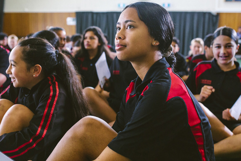 Girls in black and red school uniforms smiling and listening to a speaker in a school hall