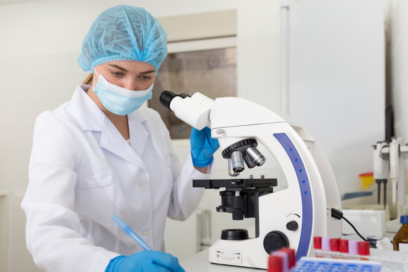 A woman in protective equipment makes notes beside a microscope 