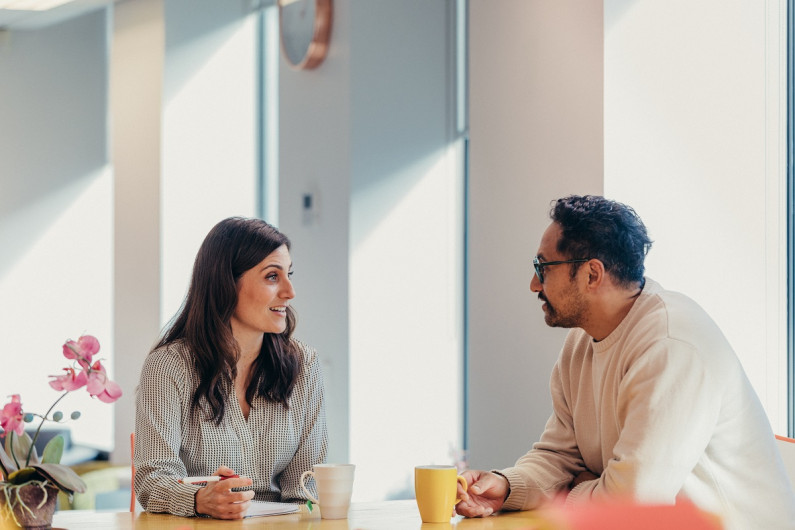 At a table in a work office, a woman with a pen and notebook talks to a man