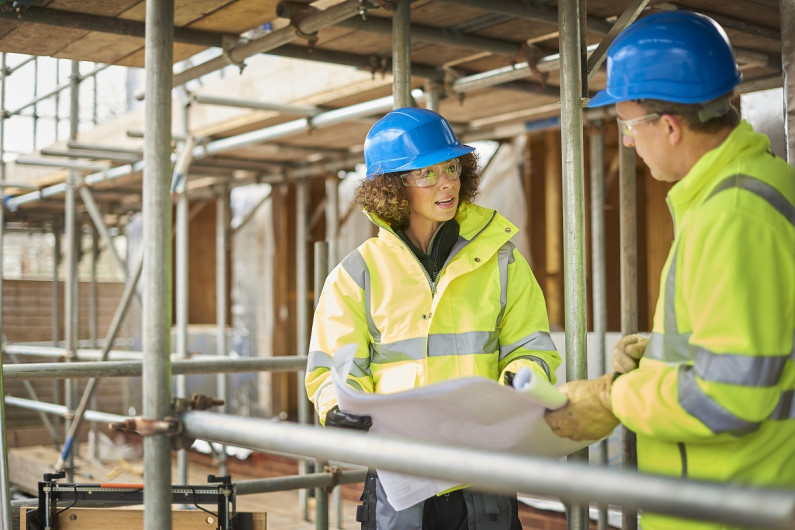 People standing on scaffolding in hard hats and yellow jackets look at a plan