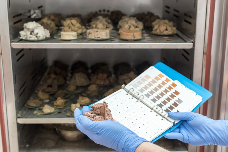 Gloved hands hold a pile of soil and a notebook full of soil colours, in front of a science fridge that contains different types of soil samples