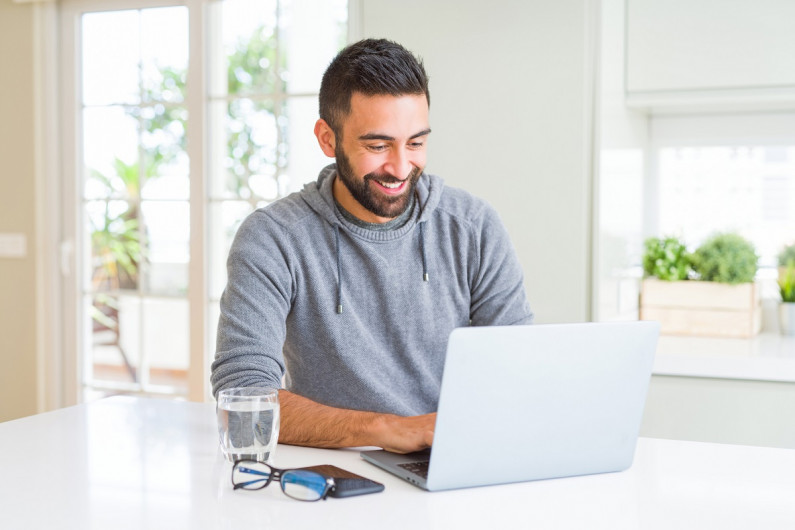 A man sits at his kitchen bench and smiles at his laptop