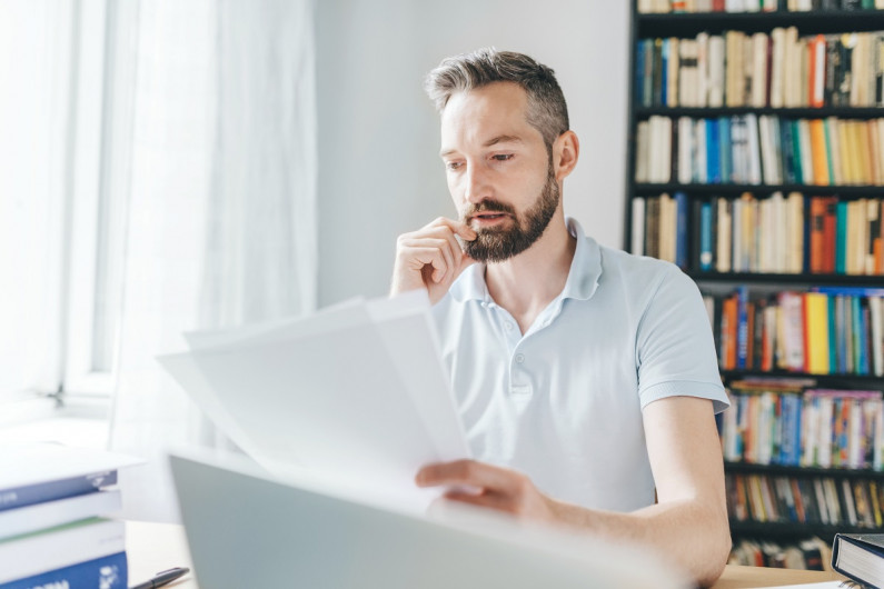 A man sits at a home desk reading sheets of paper