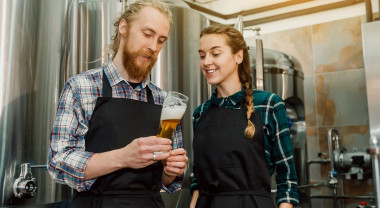 A young man and woman stand in front of beer vats inspecting a glass of beer.