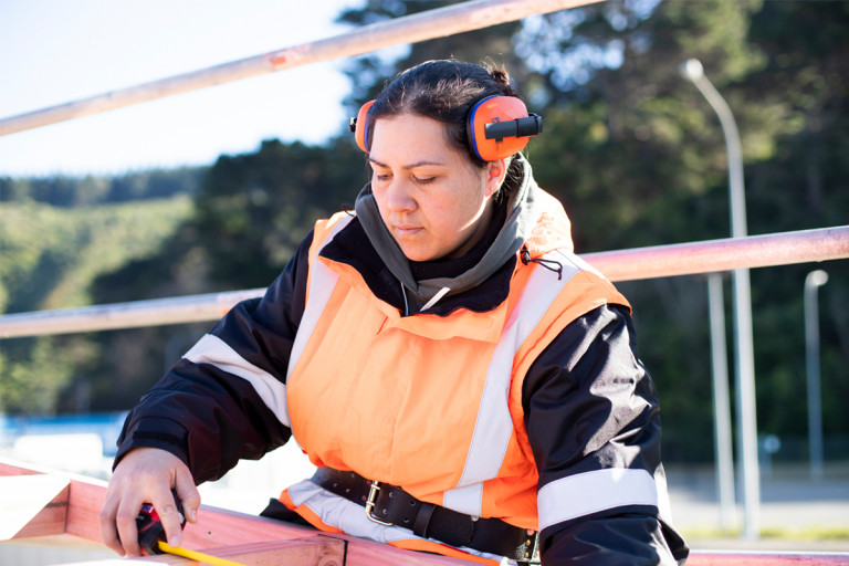 A woman wearing safety gear saws into wood on a building site
