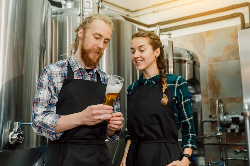 A young man and woman stand in front of beer vats inspecting a glass of beer.