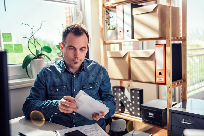 A man sits at a desk in an office full of boxes. He's putting a letter into an envelope and looking thoughtful
