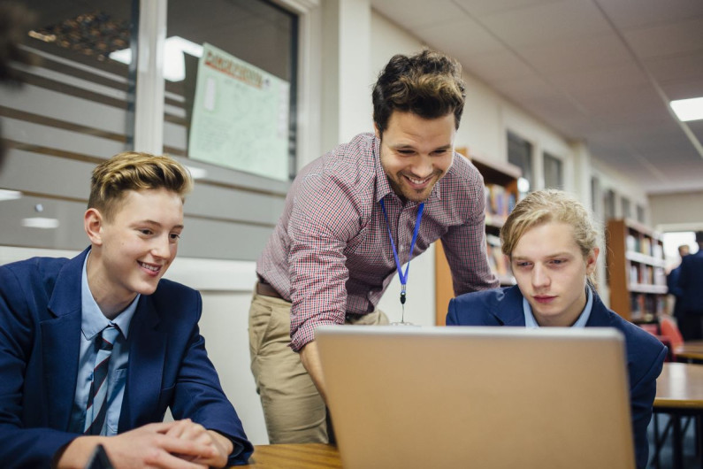 Teacher leans over a laptop two male students are looking at 