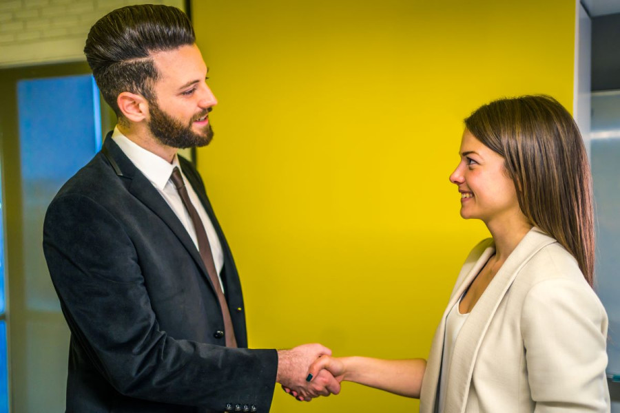 A man and a woman both dressed in suits and smiling shake hands in a modern office foyer