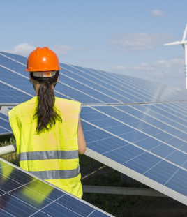 A solar panel installer sits on the roof looking at a wind turbine.