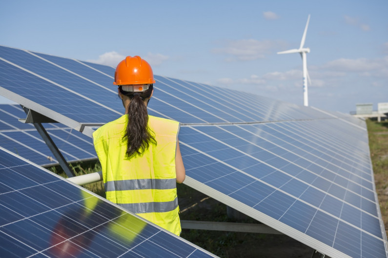 A solar panel installer sits on the roof looking at a wind turbine.