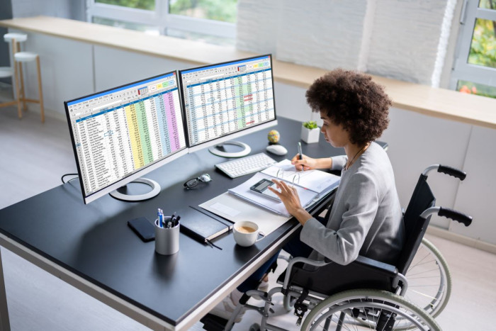 A woman in a wheelchair sits at a table looking at two large computer screens covered in spreadsheets