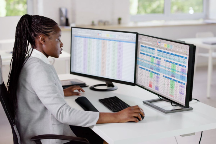 A woman in business clothes sits at a desk in front of two large computer screens showing spreadsheets