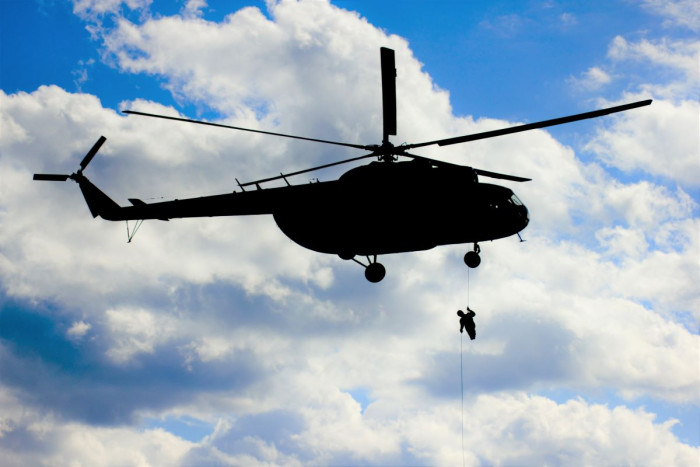 A helicopter hovers in a blue and cloudy sky with a person dangling below on a rope 