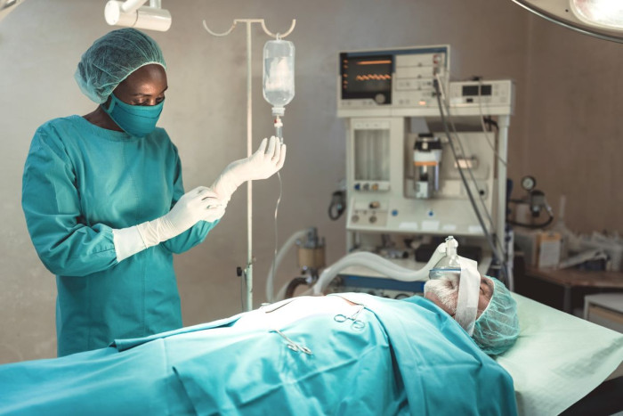 An anaesthetic technician wearing a surgical gown, mask and gloves stands beside a patient lying in an operating theatre wearing a mask, and with a monitoring machine behind him