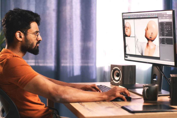 A man sits at a desk using a computer keyboard. On his computer screen are two views of a video game character he is animating 