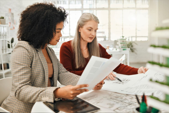 Two women sit at a desk in an office comparing documents. An architectural drawing is on the table