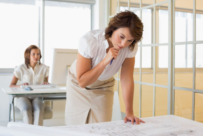 A woman in office clothes stands looking at architectural blueprints on a desk in an office. Her colleague sits at a desk further back in the office