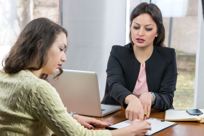 A young woman in casual clothes writes on a piece of paper where a woman in business clothes is pointing