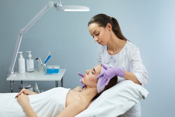 A beauty therapist wearing a white smock and purple gloves holds a clients' head in a beauty salon. The client is wearing a towel, has her hair tied back and is lying on a bed 