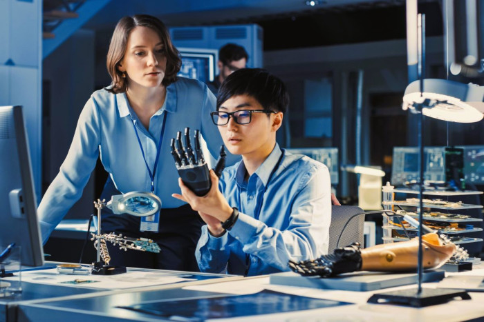 A man and a woman look at a high tech artificial hand skeleton, with a prosthesis on the desk beside them
