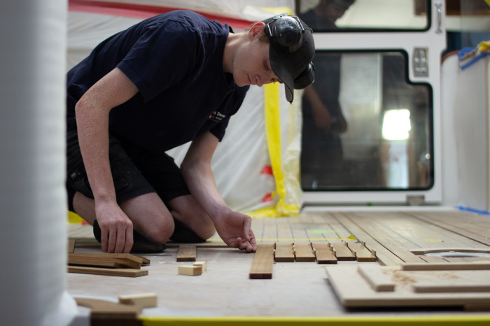 A man kneels on a floor inside a new boat, linking up wooden decking planks