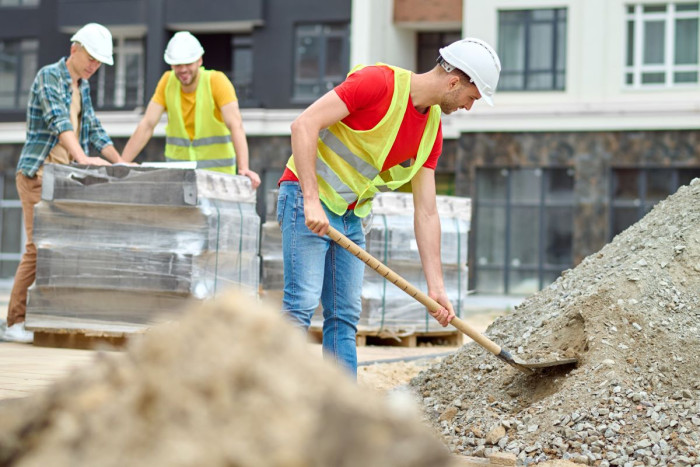 A labourer in a high viz vest and a hard hat shovels gravel while other workers look on 