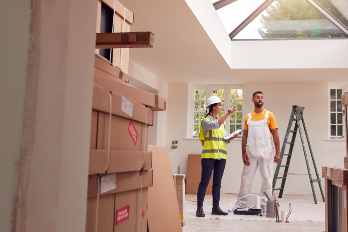 A woman in a high viz vest and hard hat holding a clip board talks to a man in painter's overalls, in a room that's under construction  