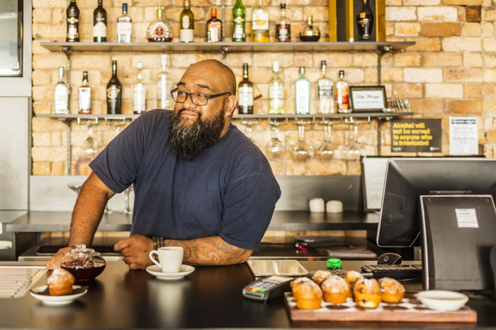  A man with a greying beard stands at the counter of a cafe with a cup of tea and a cake on a plate. Other cakes are displayed on the counter