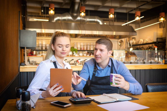 A man and a woman are sitting at a table in a cafe looking at paperwork and using a calculator