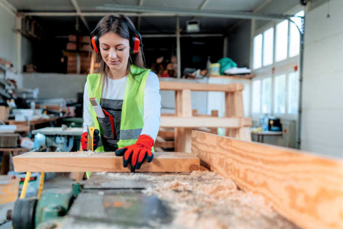 A female carpenter wearing safety glasses and a high viz vest, stands at a workbench in a workshop using an electric saw to cut a piece of wood 