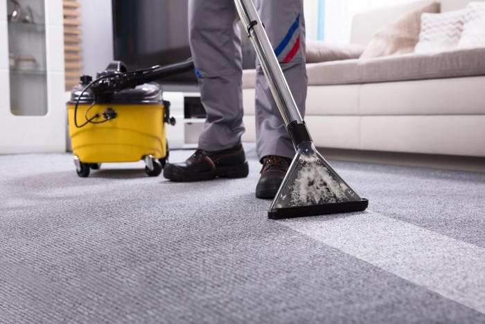 A person pulls the head of a carpet cleaner along carpeted floor, leaving a clean stripe of carpet