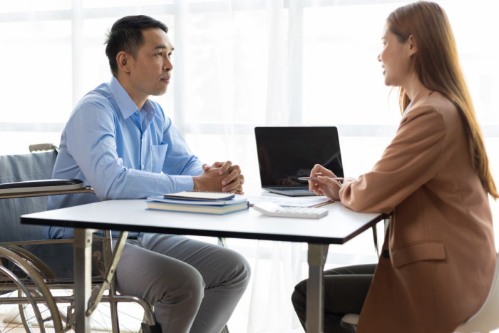 A man in office clothes in a wheelchair sits across a desk from a woman who has a laptop open 