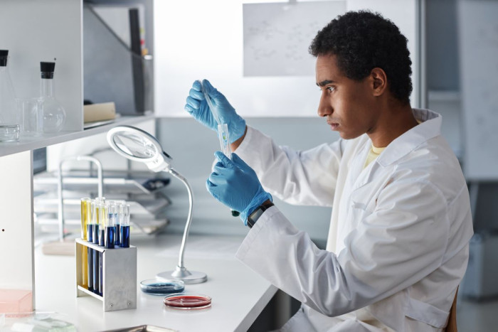 A man wearing a white lab coat and protective gloves adds liquid to a test tube 