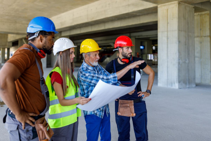 Four people wearing hard hats and other protective equipment stand in a building under construction. They're looking at paper plans and one is pointing at something outside the photograph