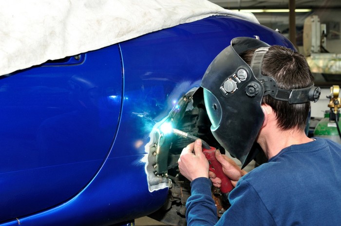 A welder wearing a protective face mask and helmet kneels down to weld a panel on a car
