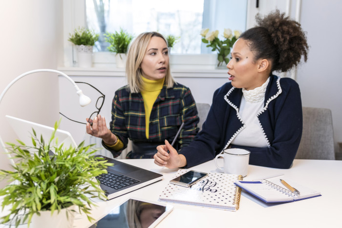 Two young women in smart office clothes talk over a computer and tablet in an office