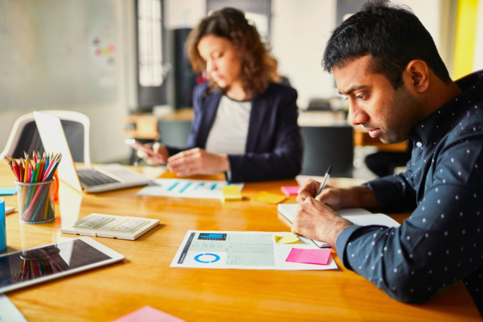 A man and woman in casual business clothes sit at a desk with laptops in front of them. She's looking at her phone, he is looking at a printed page and making notes with a pen 